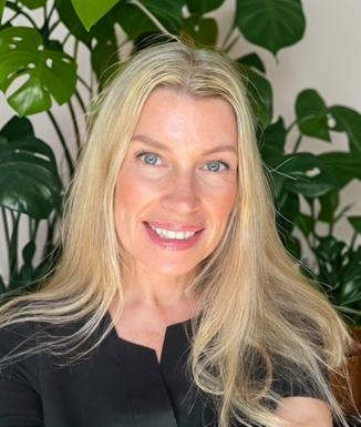 Blonde haired woman in medical uniform smiling in front of leafy plant